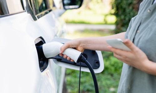 Woman with shopping bag next to a charging electric car in the yard of a country house.
